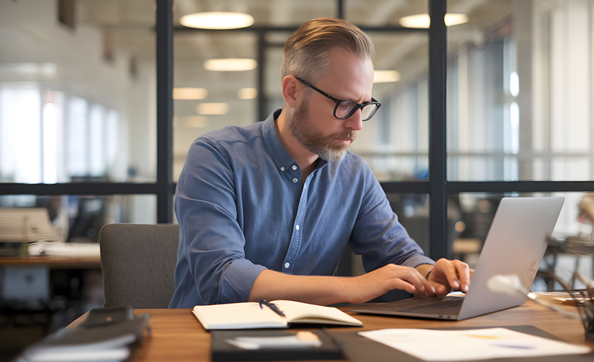 Man with glasses and beard working on a laptop in a modern office with an open notebook and papers on the desk.