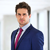 Confident businessman wearing a blue suit and red tie standing in a bright office hallway.