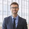 Smiling young man with glasses wearing a dark suit and tie against a blurred office window background.